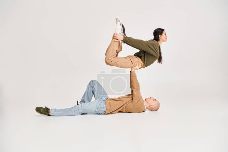 strong man in casual wear lying on floor and lifting brunette woman on grey, couple of acrobats