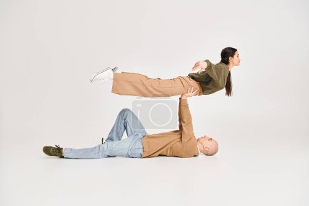 homme fort en tenue décontractée couché sur le sol et levant femme brune en studio, couple d'acrobates