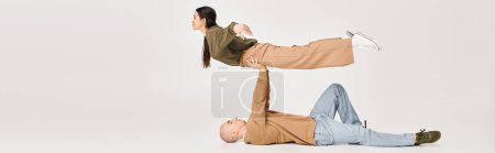 man in casual wear lying on floor and lifting brunette woman in studio, couple of acrobats banner