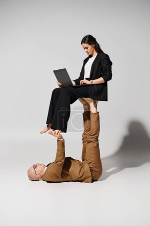 Photo for Businesswoman in attire with laptop balancing on feet of man in suit on grey, couple of acrobats - Royalty Free Image