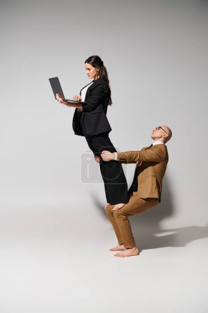 Woman in business attire with laptop balancing on laps of man in suit on grey, couple of acrobats