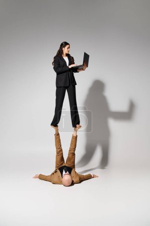 Woman in business attire with laptop balancing on feet of man formal wear, couple of acrobats
