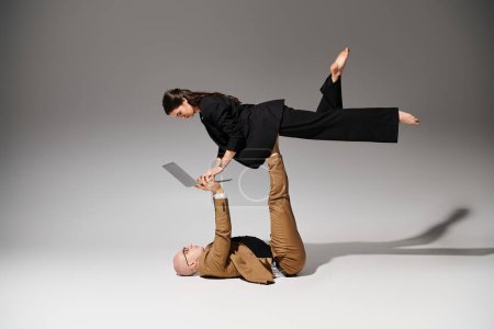 woman in business attire with laptop balancing with support of man on grey, couple of acrobats