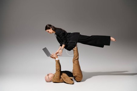 Téléchargez les photos : Femme en tenue de travail avec ordinateur portable équilibrant sur les pieds de l'homme en costume sur gris, couple d'acrobates - en image libre de droit