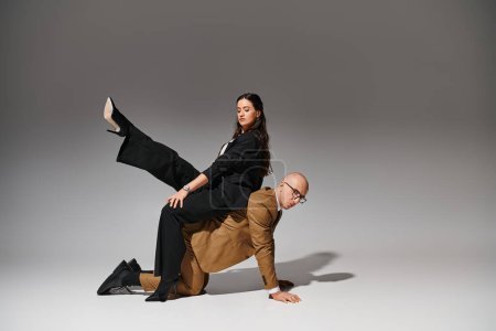 Business couple in stylish suits in creative acrobatic pose, woman sitting on back of man in studio