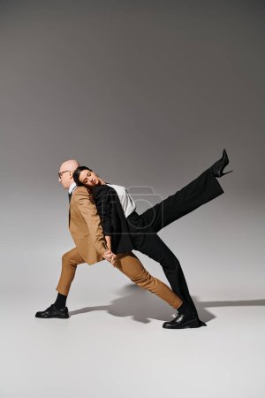 partners in coordinated dance move, woman in suit leaning on back of man in business attire on grey