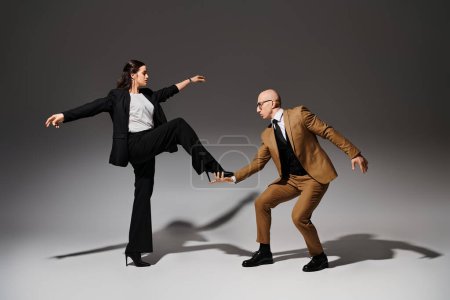 brunette woman balancing in high heels while stepping on hand of dancing partner in suit on grey