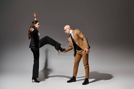 young woman balancing in high heels while stepping on hand of dancing partner in suit on grey