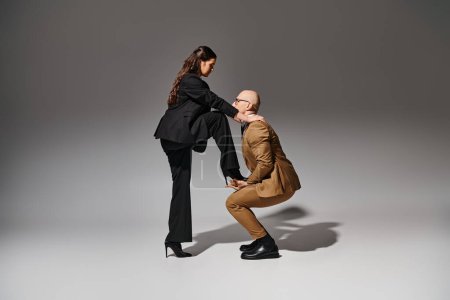 brunette woman balancing in her high heels while stepping on hand of dancing partner in suit on grey