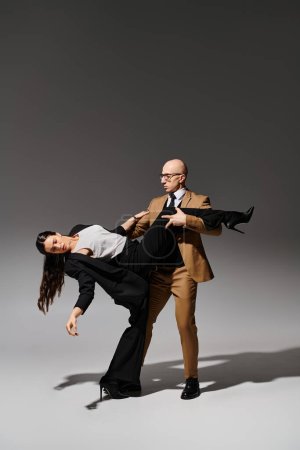 man in glasses and suit supporting leg of brunette woman in high heels during dance on grey backdrop