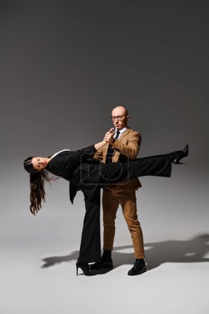 man in glasses and suit holding hand of brunette woman in high heels during dance on grey backdrop