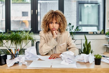 pensive curly african american businesswoman at her desk with crumpled papers, stress at work
