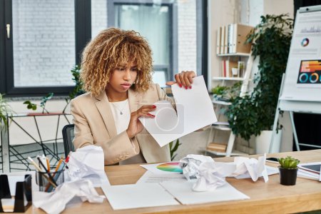 upset and curly african american businesswoman in formal wear ripping paper at her desk, stress