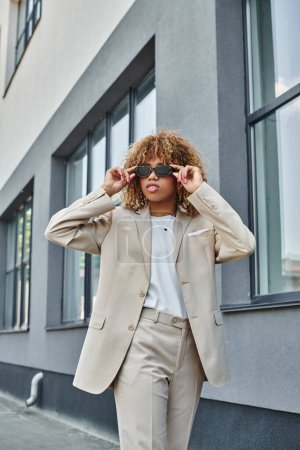curly african american woman with braces wearing sunglasses and standing near office building