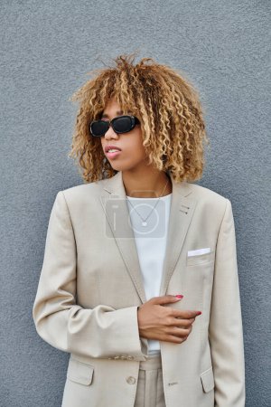 curly african american woman with braces in suit and sunglasses standing near office building