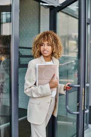 Mujer de negocios afroamericana sonriente con la carpeta de celebración de pelo rizado y salir del edificio de oficinas