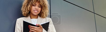 happy african american businesswoman in formal wear looking at notebook near office building, banner