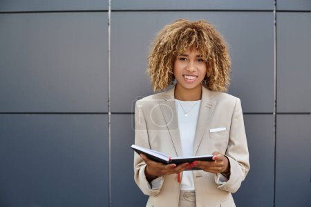 positive afrikanisch-amerikanische Geschäftsfrau in formeller Kleidung hält ihr Notizbuch in der Nähe eines Bürogebäudes