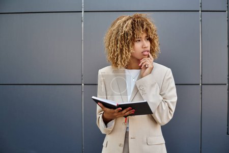 nachdenkliche afrikanisch-amerikanische Geschäftsfrau in offizieller Kleidung hält ihr Notizbuch in der Nähe eines Bürogebäudes