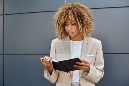pensive african american businesswoman in formal wear looking at her notebook near office building