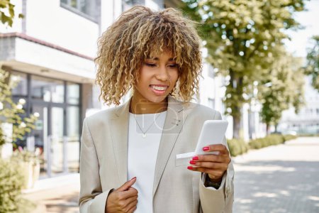 mujer de negocios afroamericana feliz en frenillos caminando con teléfono inteligente cerca del edificio de oficinas