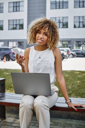 happy african american freelancer with smartphone working on her laptop on bench in a park setting
