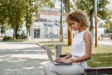 Freelancer afroamericana rizada concentrada trabajando en su computadora portátil en un banco en un entorno de parque