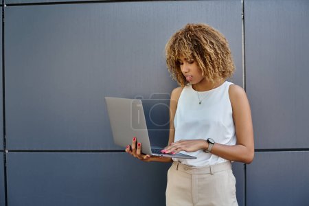 Téléchargez les photos : Femme d'affaires afro-américaine bouclée avec bretelles debout avec son ordinateur portable près du mur gris du bâtiment - en image libre de droit