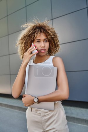 Photo for Modern young professional, african american woman with laptop making a call against grey wall - Royalty Free Image