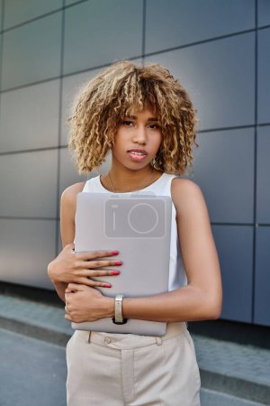 Photo for Curly african american businesswoman with braces standing with her laptop near office building - Royalty Free Image