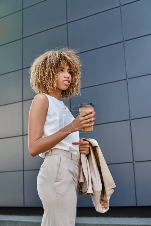 Stilvolle und lockige afrikanisch-amerikanische Geschäftsfrau mit Kaffee vor einem modernen Gebäude