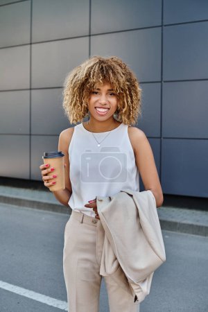 Téléchargez les photos : Heureuse et frisée femme d'affaires afro-américaine avec café debout contre un bâtiment moderne - en image libre de droit