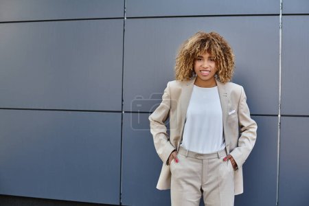 young happy black businesswoman in suit standing with hands in pockets against grey building