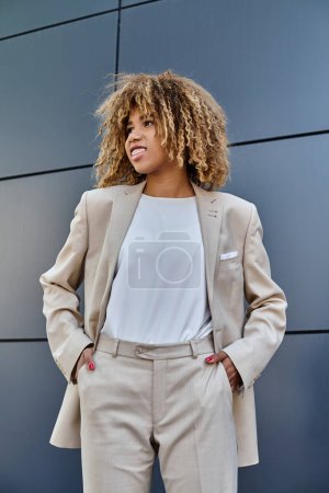 happy african american businesswoman in suit standing with hands in pockets against grey building