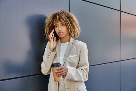 curly african american businesswoman standing with coffee and talking by phone against grey wall