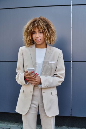 Téléchargez les photos : Heureuse femme d'affaires afro-américaine avec les cheveux bouclés debout en costume et en utilisant son smartphone - en image libre de droit