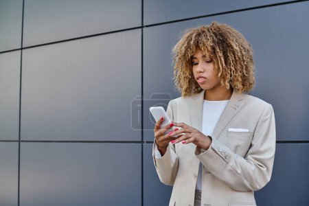 Téléchargez les photos : Jeune femme d'affaires afro-américaine avec les cheveux bouclés debout en costume et en utilisant son smartphone - en image libre de droit