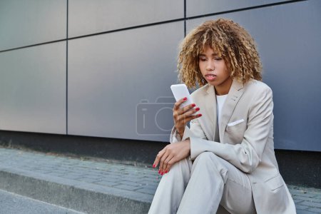 jeune femme d'affaires afro-américaine avec les cheveux bouclés debout en costume et en utilisant smartphone à l'extérieur