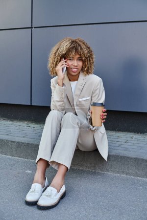 mujer afroamericana en traje sentado en parapeto con café para ir y hablar en su teléfono inteligente