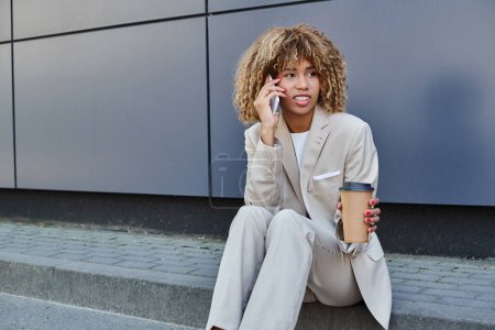 Téléchargez les photos : Jeune femme afro-américaine en costume assis sur parapet avec du café pour aller et parler sur smartphone - en image libre de droit