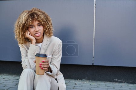 glückliche afrikanisch-amerikanische Frau im Anzug sitzt auf Brüstung mit Coffee to go und blickt in die Kamera