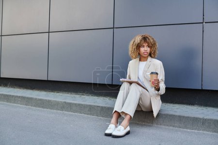 Stilvolle professionelle, afrikanisch-amerikanische Frau mit Kaffee und Ordner sitzt an der Bürowand