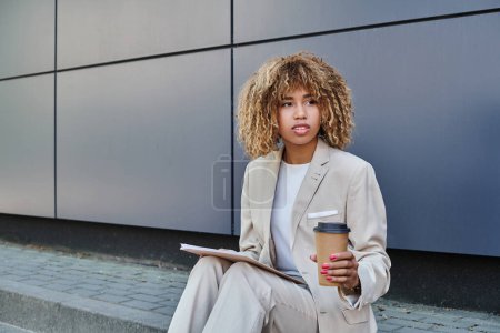 Stylish professional, african american woman with coffee and folder sitting by grey office wall
