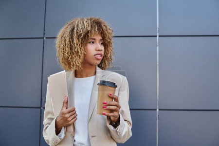 Stylish professional, african american woman with coffee and folder standing by grey office wall