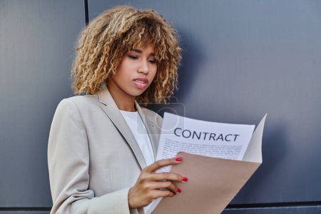 junge afrikanisch-amerikanische lockige Geschäftsfrau überprüft Vertrag unterwegs in der Nähe einer grauen Bürowand