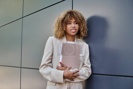 Photo for Cheerful young african american businesswoman in formal wear holding folder by grey office wall - Royalty Free Image