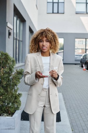 mujer de negocios afroamericana feliz en sus 20 años, de pie con una taza de café cerca del edificio de oficinas