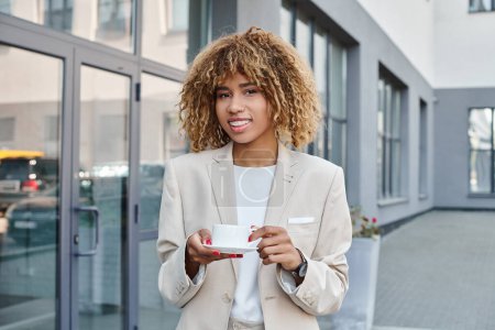 lächelnde afrikanisch-amerikanische Geschäftsfrau in ihren Zwanzigern, mit einer Tasse Kaffee in der Nähe von Bürogebäuden stehend