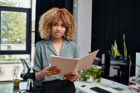 mujer de negocios afroamericana feliz en sus 20 años, de pie con café para ir y carpeta en la oficina