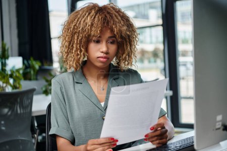 curly young african american manager reviewing document in front of computer monitor in office